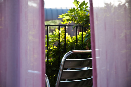 a glimpse through curtains of sun shining on a garden balcony with steel chair
